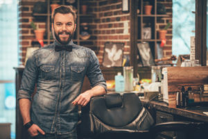 Barbershop Owner. Cheerful Young Bearded Man Looking At Camera And Holding Hand In Pocket While Leaning On Chair At Barbershop - Contabilidade em Lauro de Freitas | BM Cont