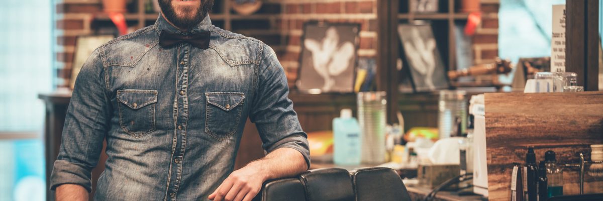 Barbershop Owner. Cheerful Young Bearded Man Looking At Camera And Holding Hand In Pocket While Leaning On Chair At Barbershop - Contabilidade em Lauro de Freitas | BM Cont